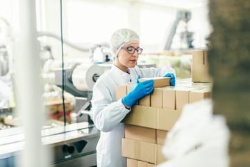 Young female employee in sterile uniform counting boxes while standing in food factory.