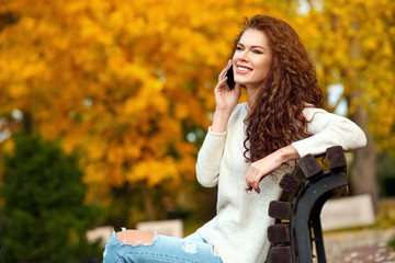 Young woman sits on a bench in the fall in a park and talks on a mobile phone