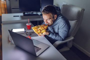 Young Caucasian architect eating pizza at office. On desk laptop. Late night work concept.