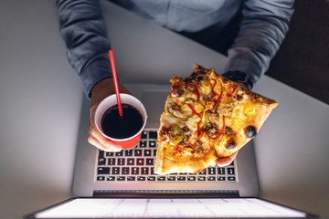 Top view of Caucasian hardworking man holding pizza and cup with juice above laptop. Late night...