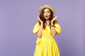 Shocked young woman in yellow dress, summer hat keeping mouth open, spreading hands isolated on pastel violet wall background in studio. People sincere emotions, lifestyle concept. Mock up copy space.