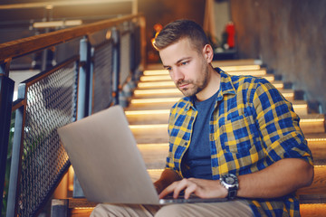Serious Caucasian handsome freelancer in plaid shirt using laptop while sitting on the stairs in cafe.