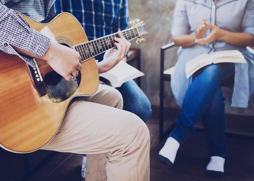 Group of  man and woman friends sitting on wooden chair while praise and worship God  by playing guitar and sing a song together in home office, Christian background small fellowship meeting concept.