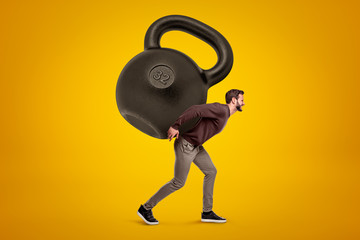 Young man in casual clothes carrying a big kettlebell on his back on yellow background