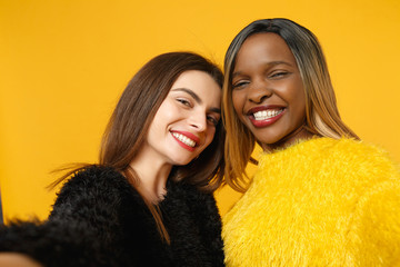 Two women friends european and african american in black yellow clothes doing selfie shot posing isolated on bright orange wall background studio portrait. People lifestyle concept. Mock up copy space