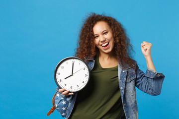 Young african american girl teen student in denim clothes, backpack hold clock isolated on blue...