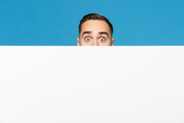Fun young man hiding, look out eyes big white empty blank billboard for promotional content isolated on blue wall background studio portrait. People emotions lifestyle concept. Mock up copy space.