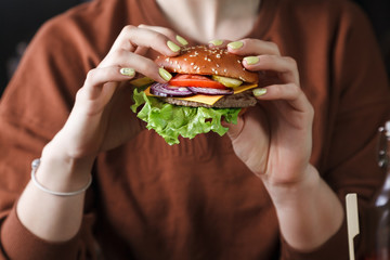 The girl is holding a delicious Burger - a fresh sesame bun, a juicy cutlet, green lettuce leaves, onions, a juicy tomato and a slice of cheese