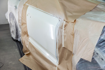 A large white car is completely covered in paper and adhesive tape to protect against splash during painting door after an accident in a workshop for body repair of vehicles with bright lighting