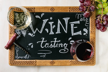 Variety of wine. Red and white wine in old fashion glasses, corkscrew, bunches of grapes, decorative chalkboard with handwritten chalk lettering Wine tasting. White marble background. Flat lay, space