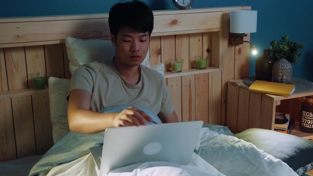 Funny attractive young Asian man in bed using gomer simpson�s hand for switching channel on tv via remote control, smiling happily. Good mood, positive emotions, having fun. Telecommunication concept