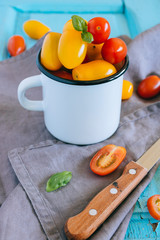 Red and yellow small cherry tomatoes, and green basil in an enameled mug on a blue background