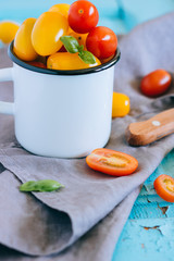 Red and yellow small cherry tomatoes, and green basil in an enameled mug on a blue background