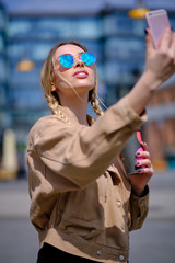 Young happy lady in a spring vacation, walking in the city, drinking coffee, photographing herself on a sunny day