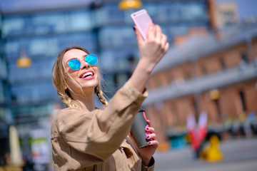 Young happy lady in a spring vacation, walking in the city, drinking coffee, photographing herself on a sunny day