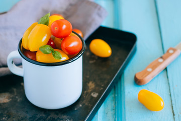 Red and yellow small cherry tomatoes, and green basil in an enamel mug on a black tray