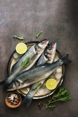 Raw sea fish sea bass on a plate with ice, rosemary and lemon, dark brown rustic background. Top view, flat lay, copy space.