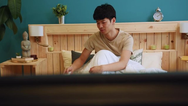 Young chinese man watches tv in bed, switches off the television via the remote control and lays down, covers himself with blanket, trying to sleep. Day end, sleeping time, dreaming. Feeling tired