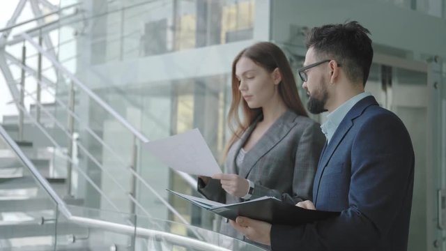Two young successful business people in suits are discussing a project in a bright modern building in high-tech style, standing near the railing. A man points his hand at an object in the distance.