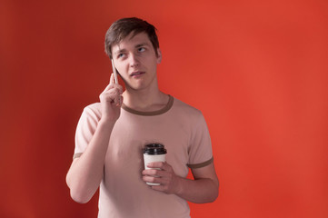 annoyed young man in beige t shirt standing and holding paper cap with coffee and talking on smartphone on orange background with copy space