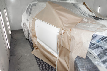 A large white car is completely covered in paper and adhesive tape to protect against splash during painting and repair after an accident in a workshop for body repair of vehicles with bright lighting