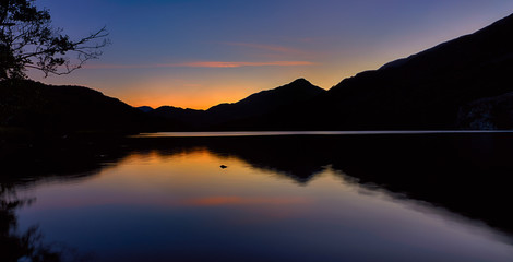 The sun has set behind the mountains around Llyn Gwynant, a lake in Snowdonia (Eryri), Wales (Cymru), UK. Silhouetting the mountains, casting a reflection off of the water.