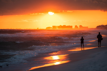 Fototapeta na wymiar Young couple watching dramatic orange red sunset in Santa Rosa Beach, Florida with Pensacola coastline coast skyline in panhandle with ocean gulf of mexico waves