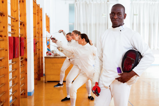 Active african american male fencer in uniform standing with mask and foil at fencing room - Powered by Adobe