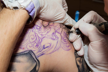 Tattoo making process over the sketch drawn by pen on male's shoulder. Tattoo machine in the hands in white gloves and new drawing making. Tattooed skin of young man stuffing a new tattoo in studio.