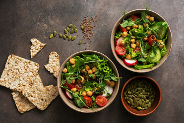 Bowls healthy vegetarian vegan salad,chickpea, lettuce, tomato, microgreen, radish, cucumber, flax seeds and pumpkin seeds with whole grain crispbreads on a dark brown background. Top view, flat lay