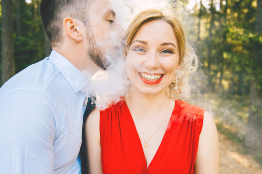 Smoking man in business clothes kissing young blonde girl with red lips, dress with decolette.  Clouds of thick smoke. Couple of lovers outdoor.  Smiling and laughing woman. E-cigarette.