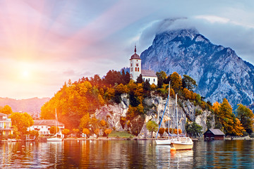 Beautiful scenic sunset over Austrian alps lake. Boats, yachts in the sunlight infront of church on the rock with clouds over Traunstein mountain at the alps lake near Hallstatt Salzkammergut Austria - 265520996