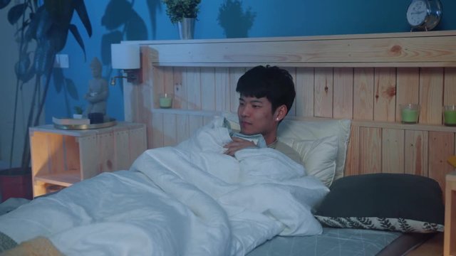 Terrified young asian man lying in bed, watching scary movies, hides under the blanket. Coziness and comfort, safe place. Entertainment concept. End of the day, relaxation, dreaming