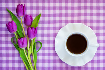 White porcelain cup of tea or coffee on saucer with wavy edge and a bouquet of purple tulips on a checkered tablecloth. Flat lay greeting card.