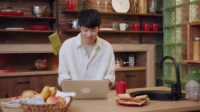 Good looking positive young Asian man actively using laptop for making video call in modern light kitchen, talking and smiling. Internet call, surfing, call app, devices and gadgets concept