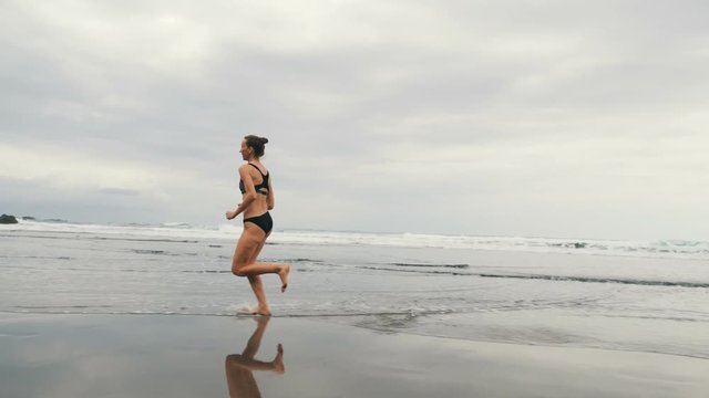 Tracking shot of attractive young woman jogging on stormy Benijo beach in Tenerife, Canary Islands. Female runner in cloudy seaside background