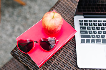 Stylish red sunglasses lying on book next to apple and white laptop with blur background. Close-up concept photo of freelance outdoor workplace in the cafe. Distant study, busy brunch, freelance job