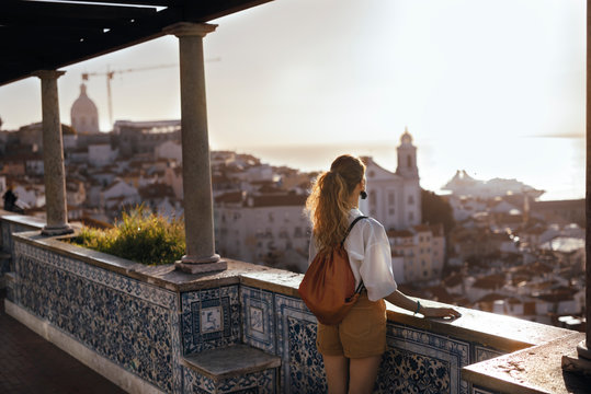 Blonde woman standing on the balcony and looking at coast view of the southern european city with sea during the sunset, wearing hat, cork bag, safari shorts and white shirt