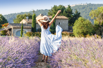 Lovely girl walking by blooming lavender fields in Luberon area in Provence, France. Beautiful girl dressing straw hat and blue boho chic dress - traditional Provencal style.