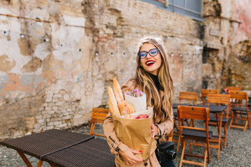 Fototapeta premium Attractive young woman came to outdoor cafe after food shopping and looks away. Stylish fair-haired girl in big glasses posing in front of old wall holding bakery bag and bouquet of purple flowers.