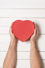 Male hands hold a red heart shaped box against the background of a white wooden table