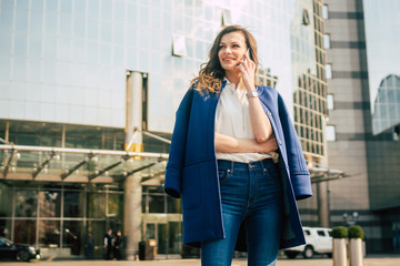 Caucasian business woman speaking by phone. Waist up portrait of a successful European business woman woman, talking on the phone, standing on glass background, modern office building. Sunny weather