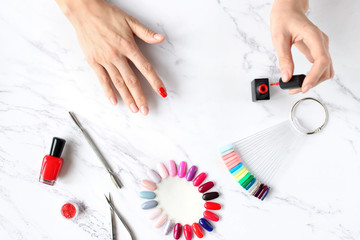 Beautiful woman hands painting nails with red nail polish on marble table with manicure set on it, top view.