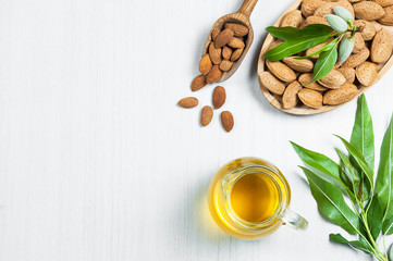 Top view Glass bottle of Almond oil and almond nuts in wooden shovel, almonds with shell in bamboo bowl on white table with green fresh raw almonds on almond tree branch