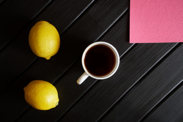 A white cup with black tea stands on a black table next to two yellow lemons and a pink napkin....