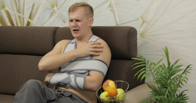 Painful man with a broken arm wearing arm brace sitting on a sofa eating apple