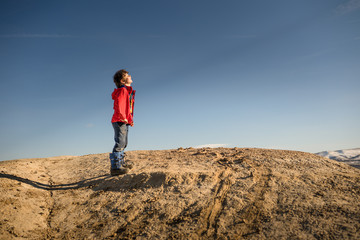 Fototapeta premium Young boy in red jacket facing sun on top of mountain with blue sky