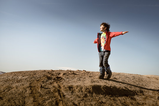 Young boy in red jacket on mountain facing sun with arms outstretched and hair blowing