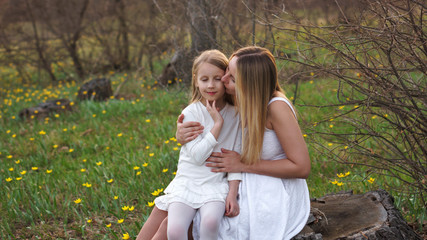 Attractive mother gently kisses her daughter on her lap. Family on a walk in spring forest on a flowery meadow. Girl enjoys a tender moment. Concept of mom love, affection and care. Mother's Day