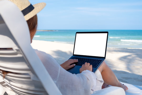 Mockup image of a woman using and typing on laptop computer with blank desktop screen while laying down on beach chair on the beach